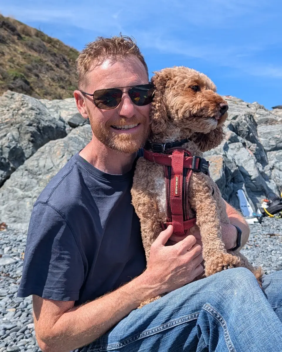 Andy sitting on a pebble beach with his dog Ruby in his arms